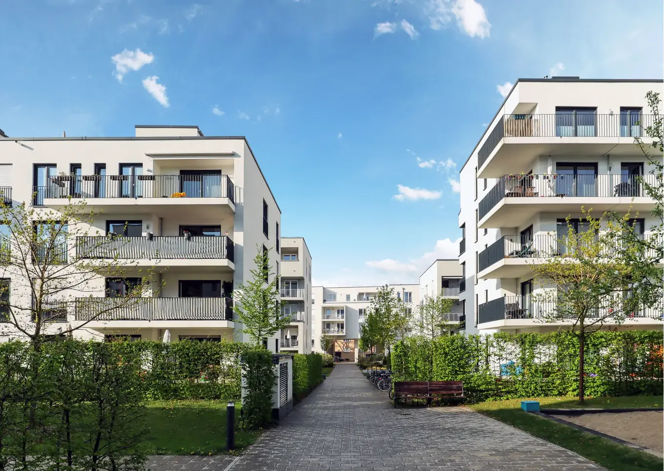 Twee moderne appartementsgebouwen met balkons langs een gemeenschappelijk aangelegd wandelpad met groen.