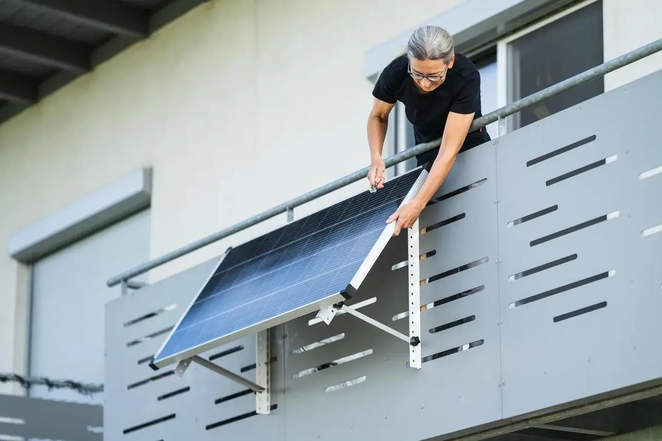 Vrouw bevestigt een zonnepaneel met aluminium montagebeugel aan de buitenzijde van een grijze balkonafsluiting van een appartement.
