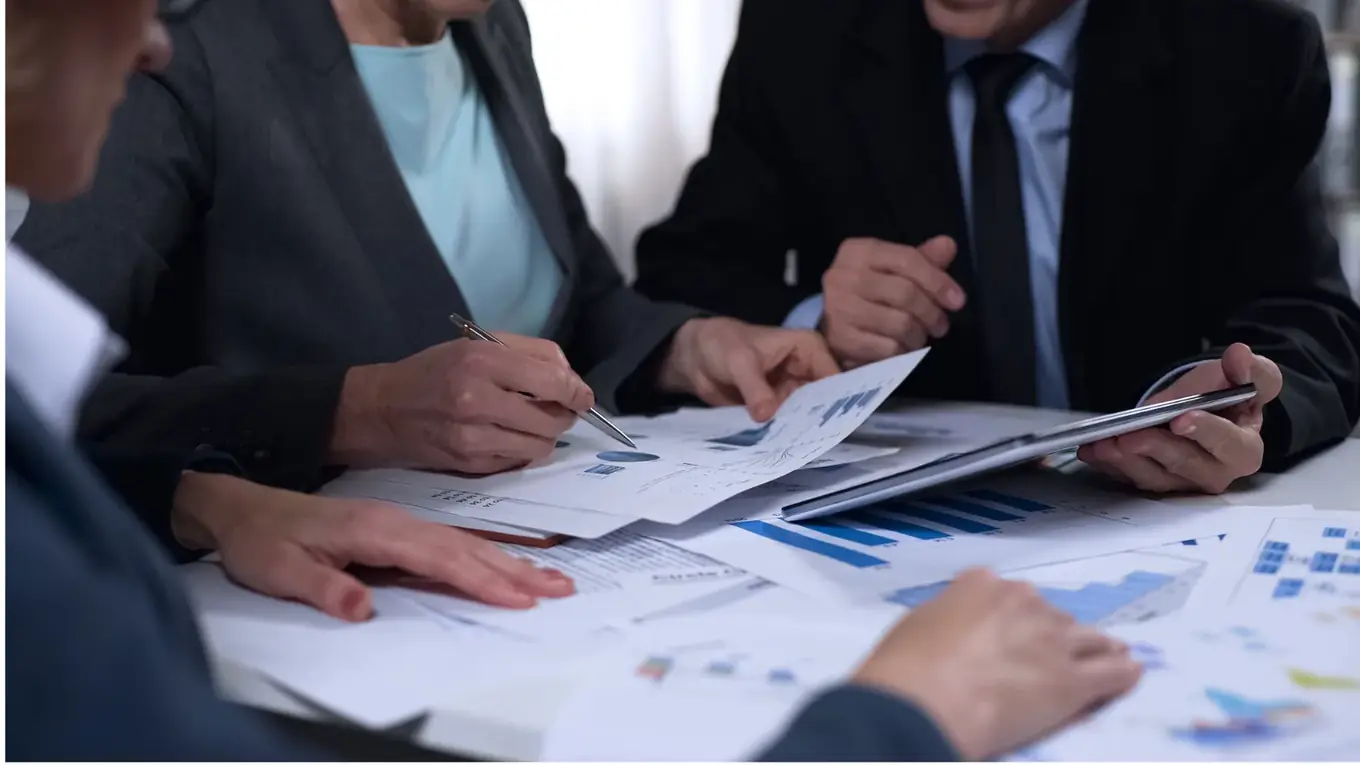 Drie mensen in pak overleggen aan een tafel vol documenten met staafdiagrammen, één wijst met een pen aan en een ander houdt een tablet vast