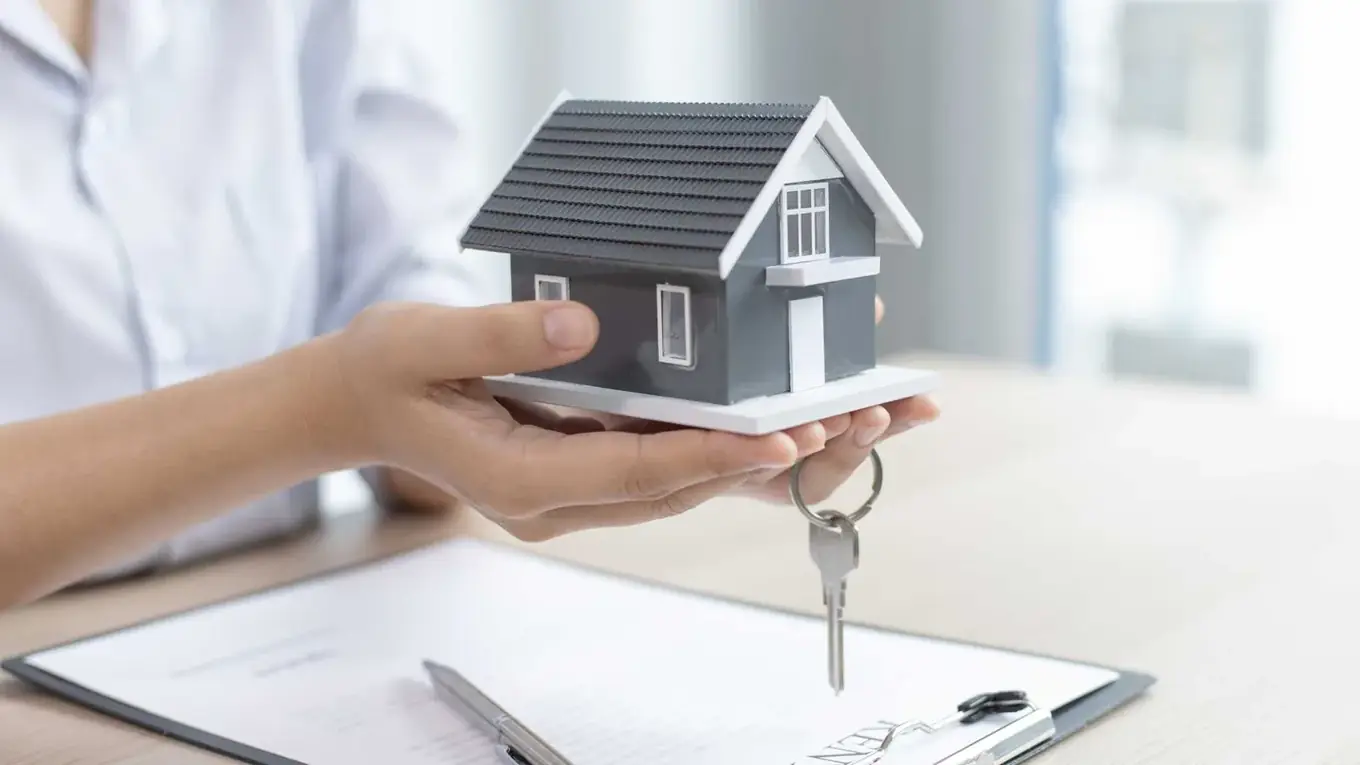 Hand houdt een klein grijs-wit modelhuis vast met een sleutel eraan, boven een klembord met een document en een pen
